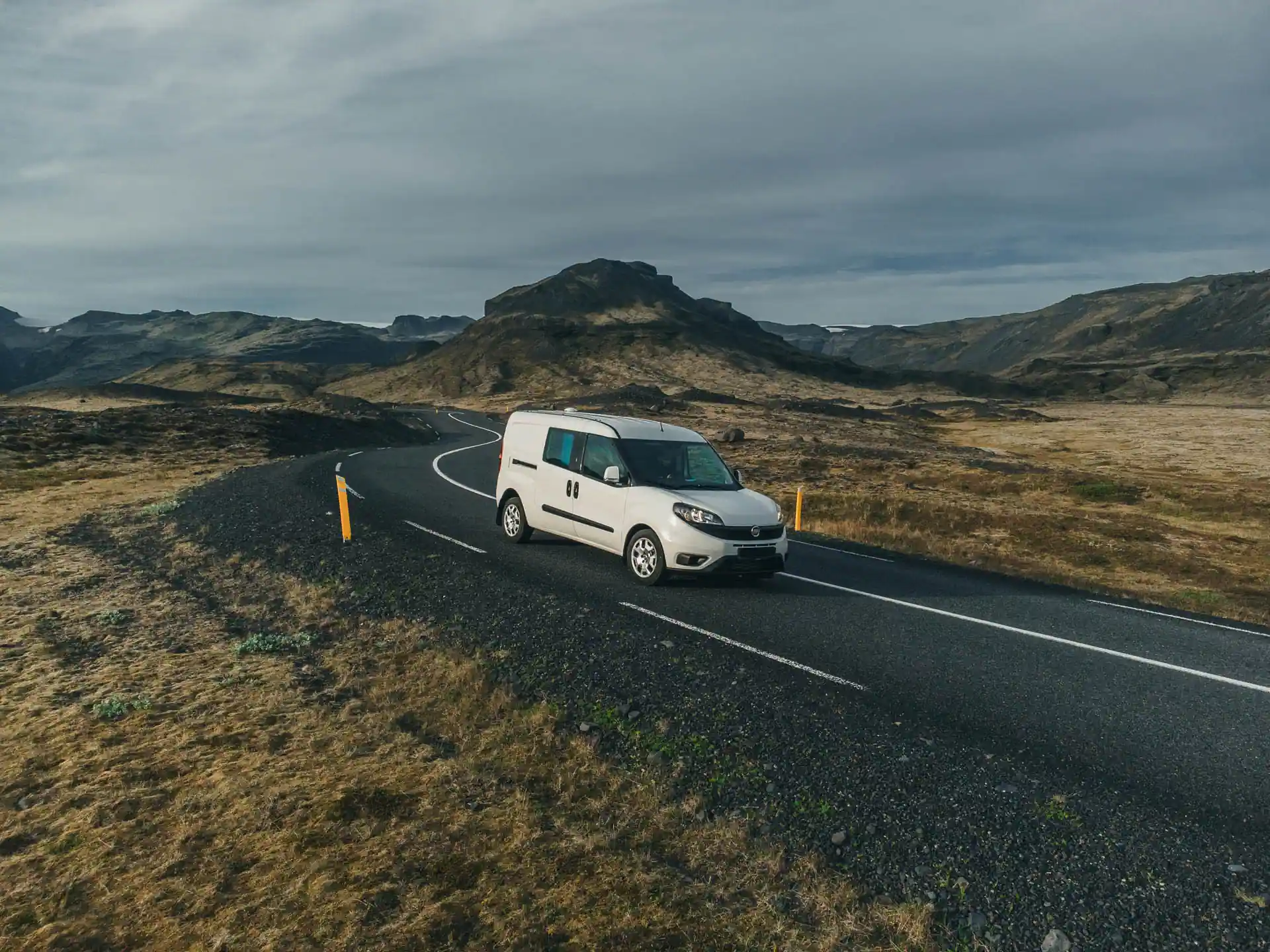 White Fiat Doblo campervan driving along winding Icelandic road with mountains and moody cloudy sky in the background