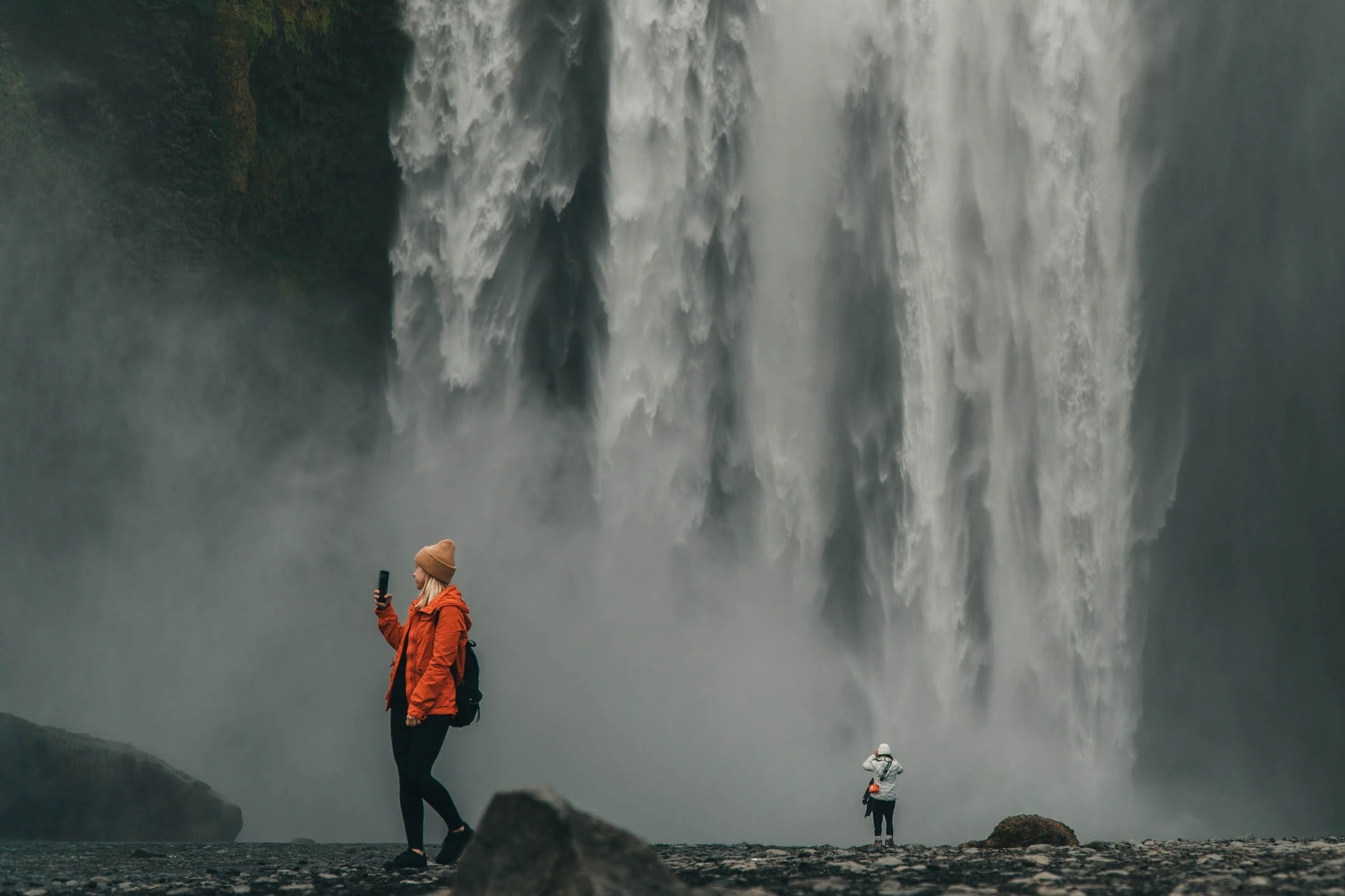 Traveler in an orange jacket taking photos at Skógafoss waterfall on an Iceland Ring Road campervan trip