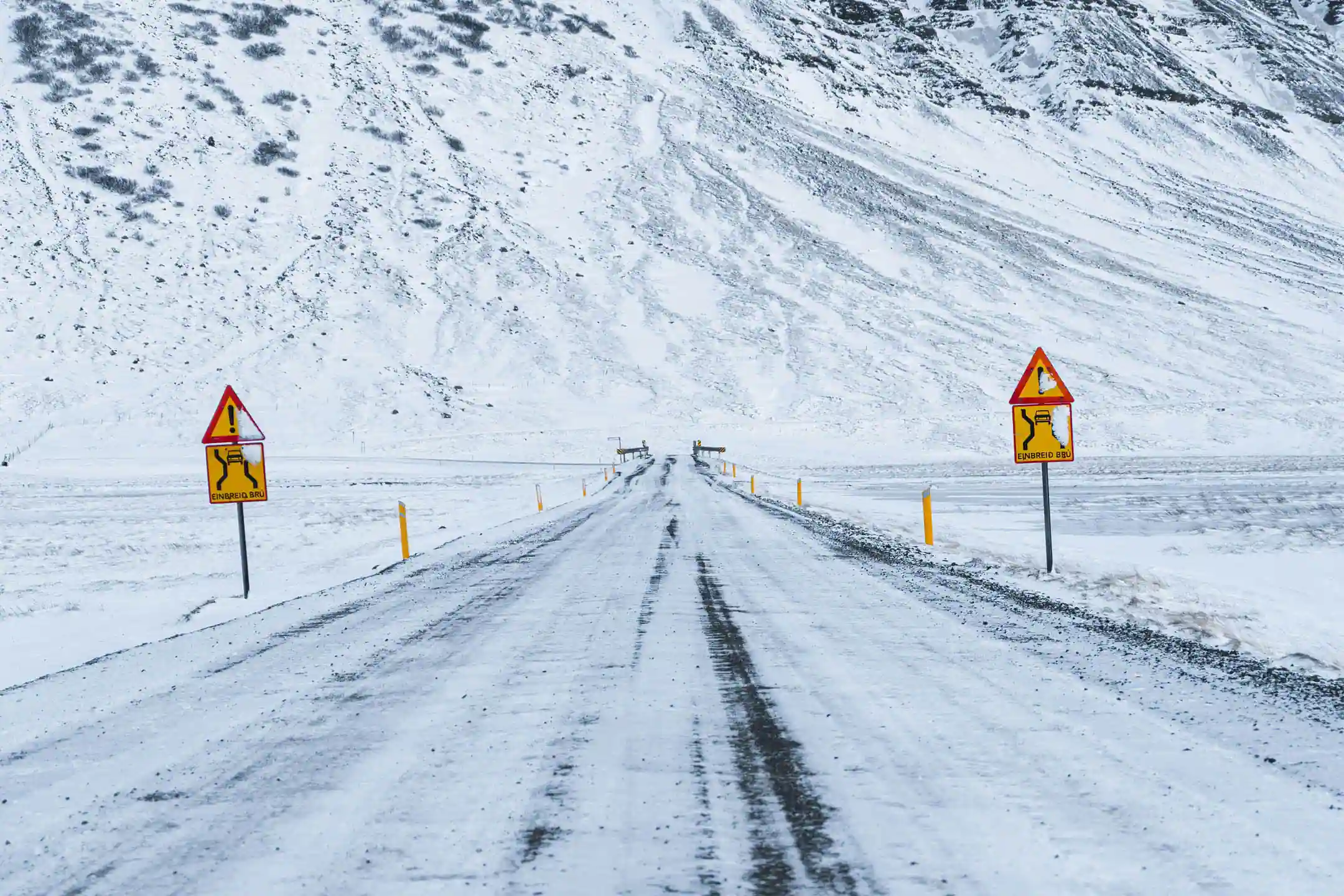 Snowy winter road in Iceland with yellow warning signs for a single-lane bridge and a sharp turn ahead