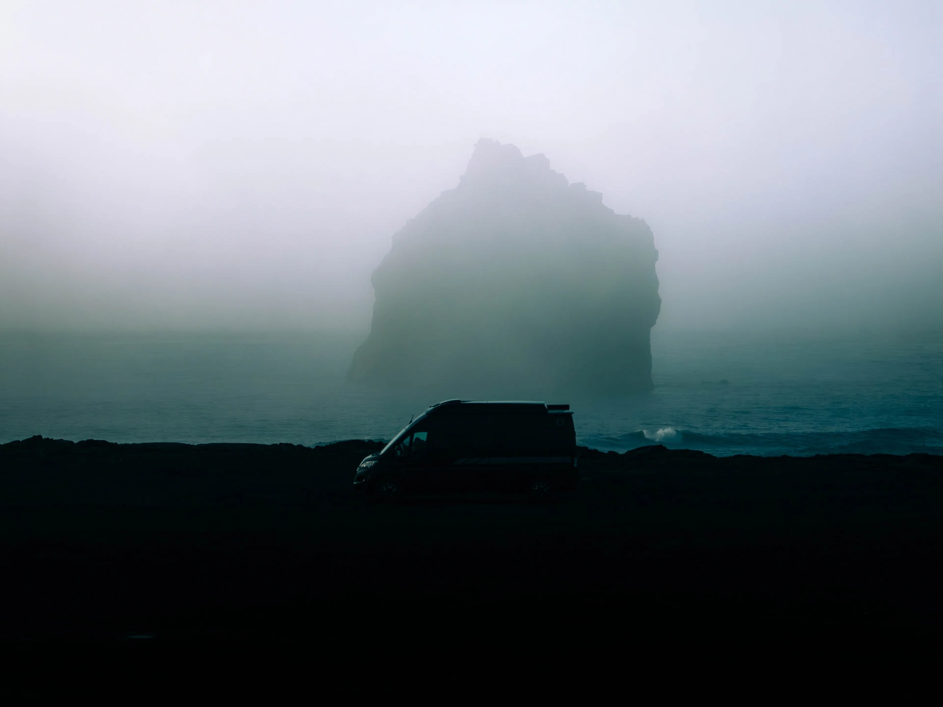Campervan parked on a dark Icelandic coastline with sea mist and rock formations, showing the appeal of a flexible eclipse base camp.