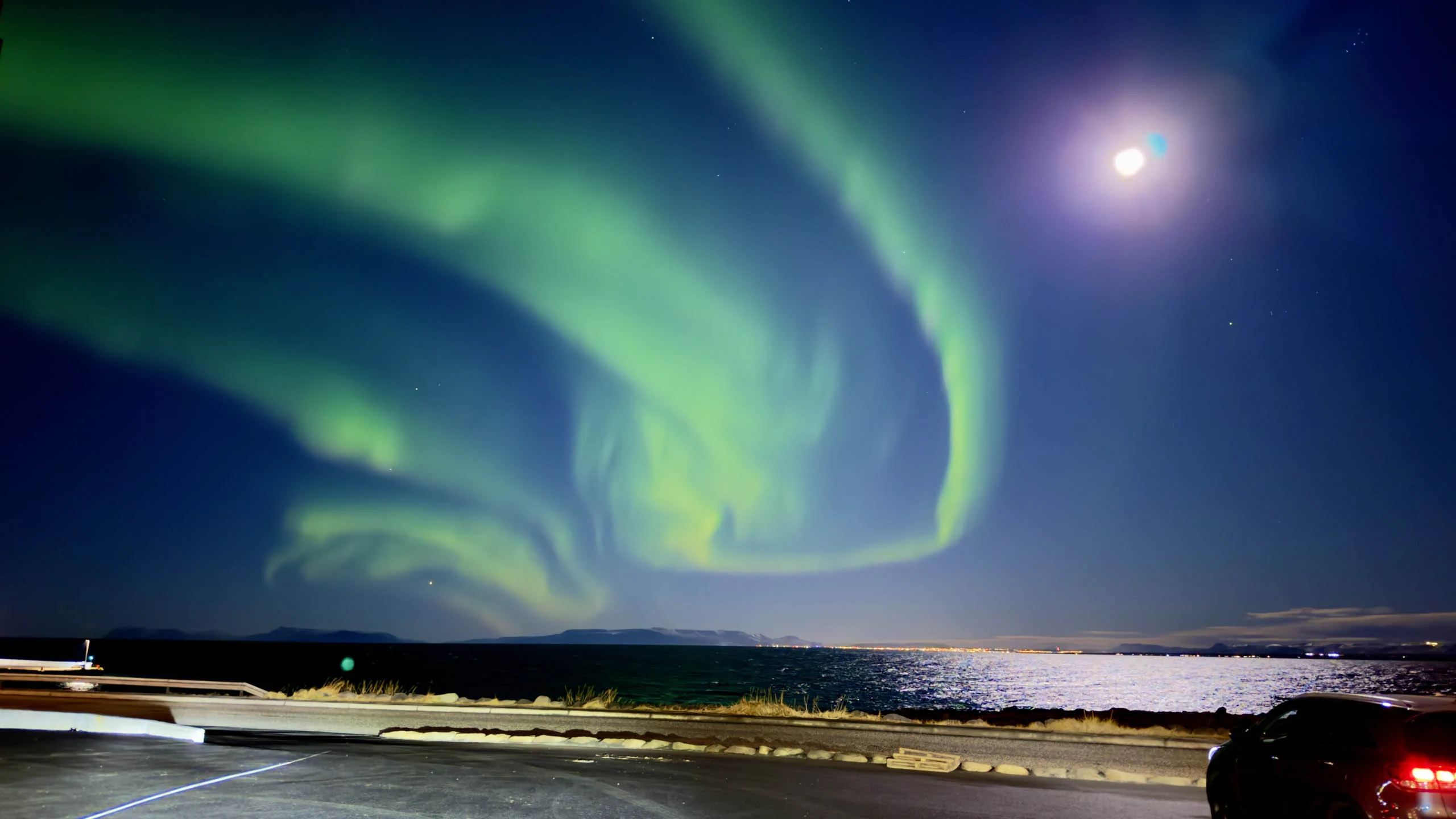 Intense green and red Northern Lights in Iceland illuminating the night sky above travelers parked along a dark roadside.