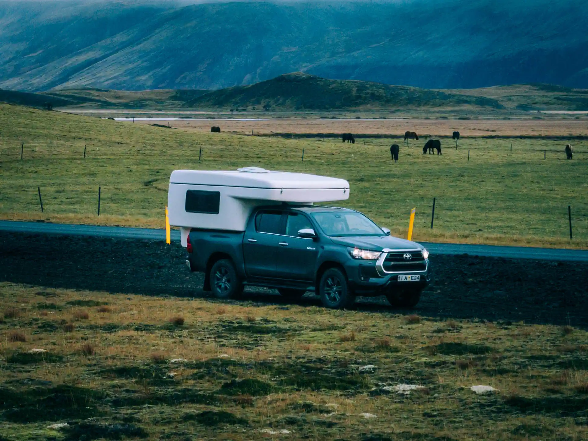A Toyota Hilux campervan parked in a wide Icelandic valley with Icelandic horses grazing in the background. 