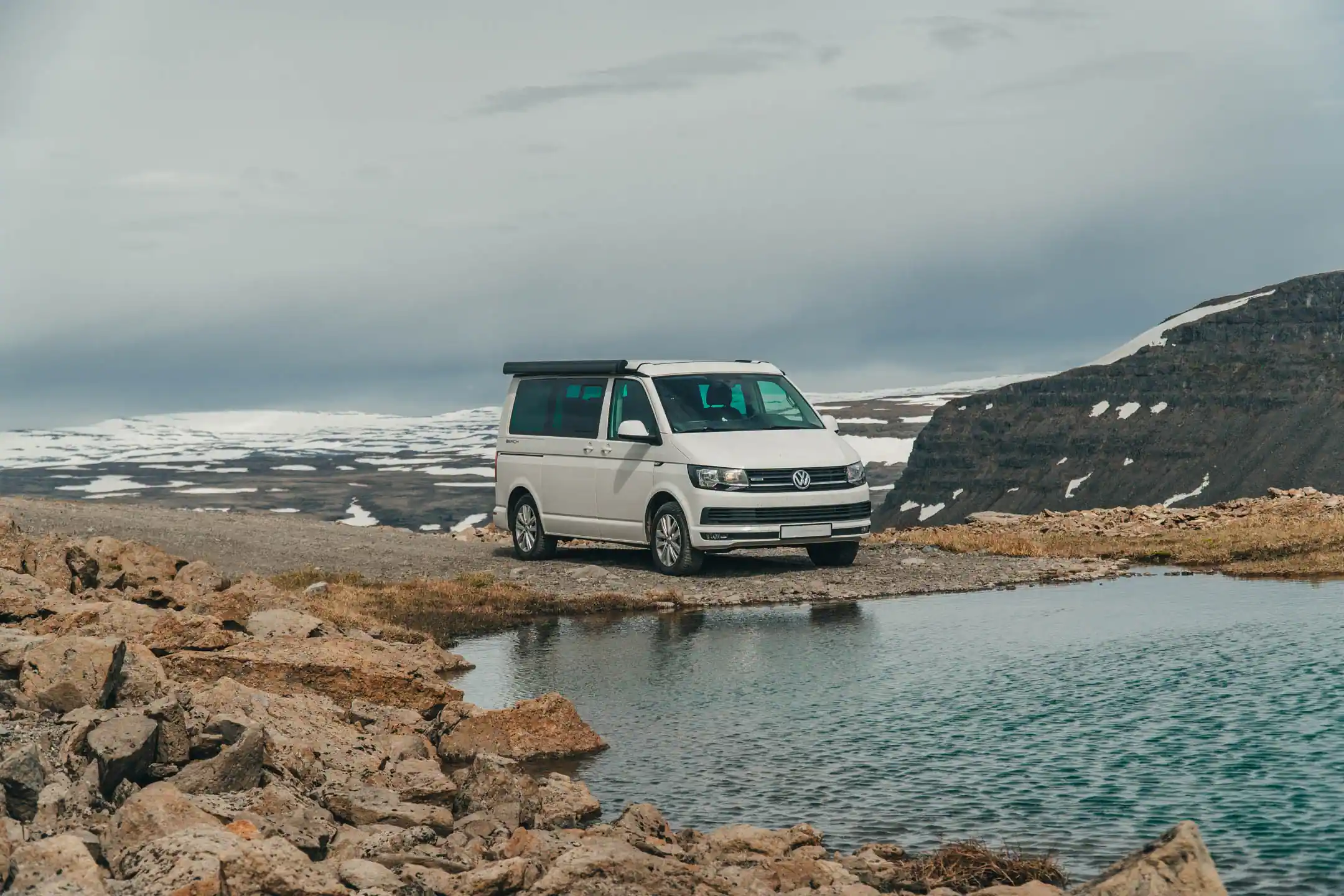 VW campervan parked beside a shallow river in Iceland with snowy mountains in the background, illustrating safe preparation before a river crossing