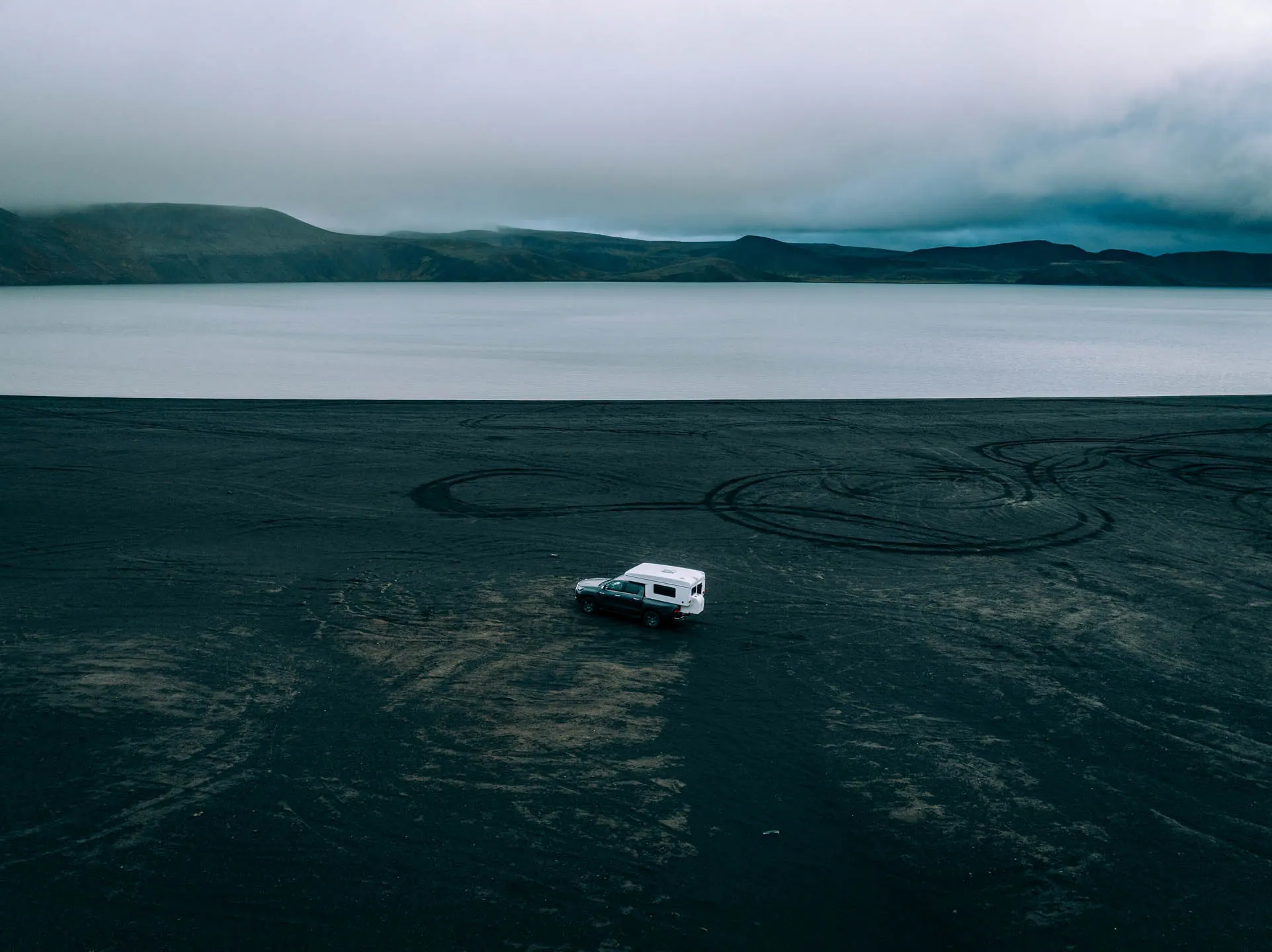 4x4 pickup camper driving across a black volcanic beach beside a calm lake with distant mountains under an overcast sky in Iceland