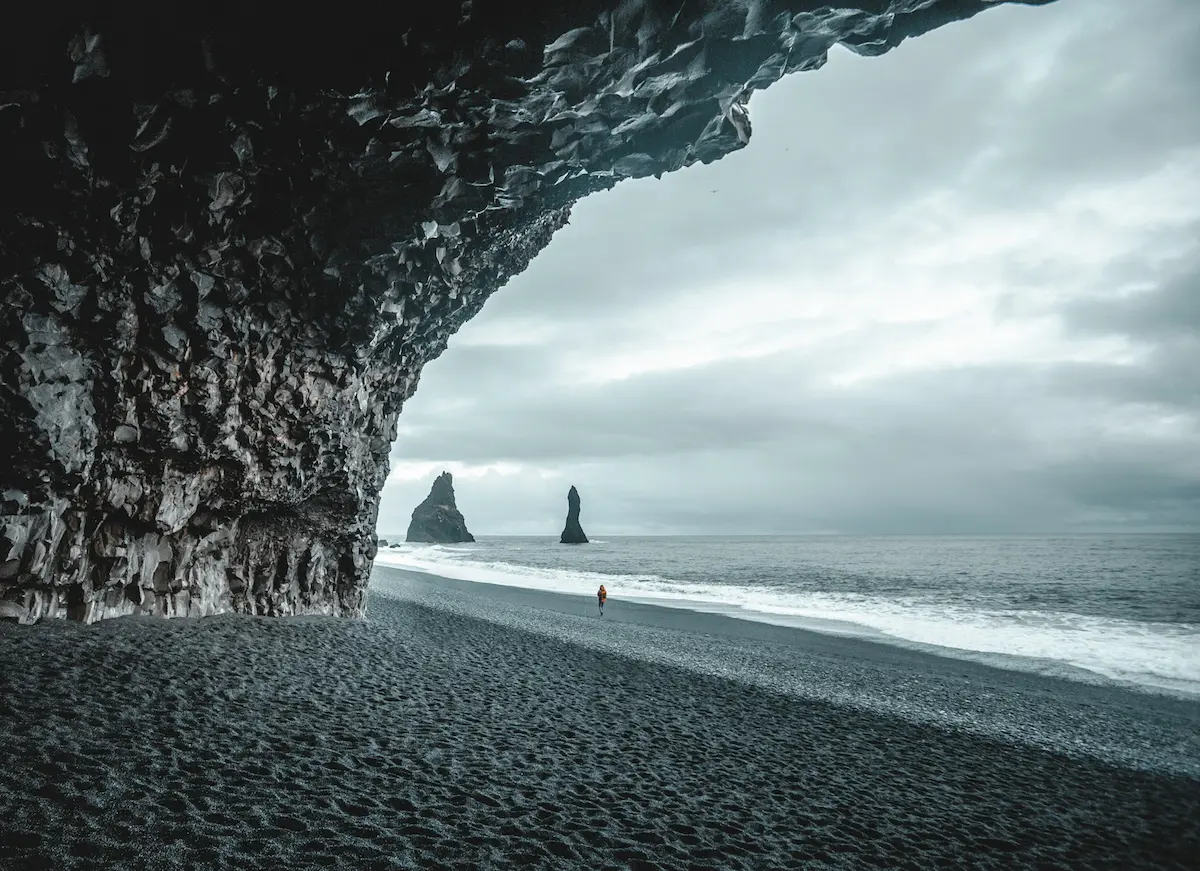 View from inside the basalt cave at Reynisfjara Black Sand Beach, looking out toward the Reynisdrangar sea stacks and the Atlantic Ocean under cloudy skies.